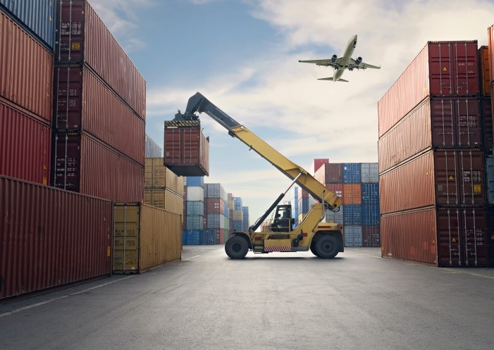 Umschlag in der Logistik: Flugzeug fliegt über Containerhafen, an dem ein Container umgeschlagen wird